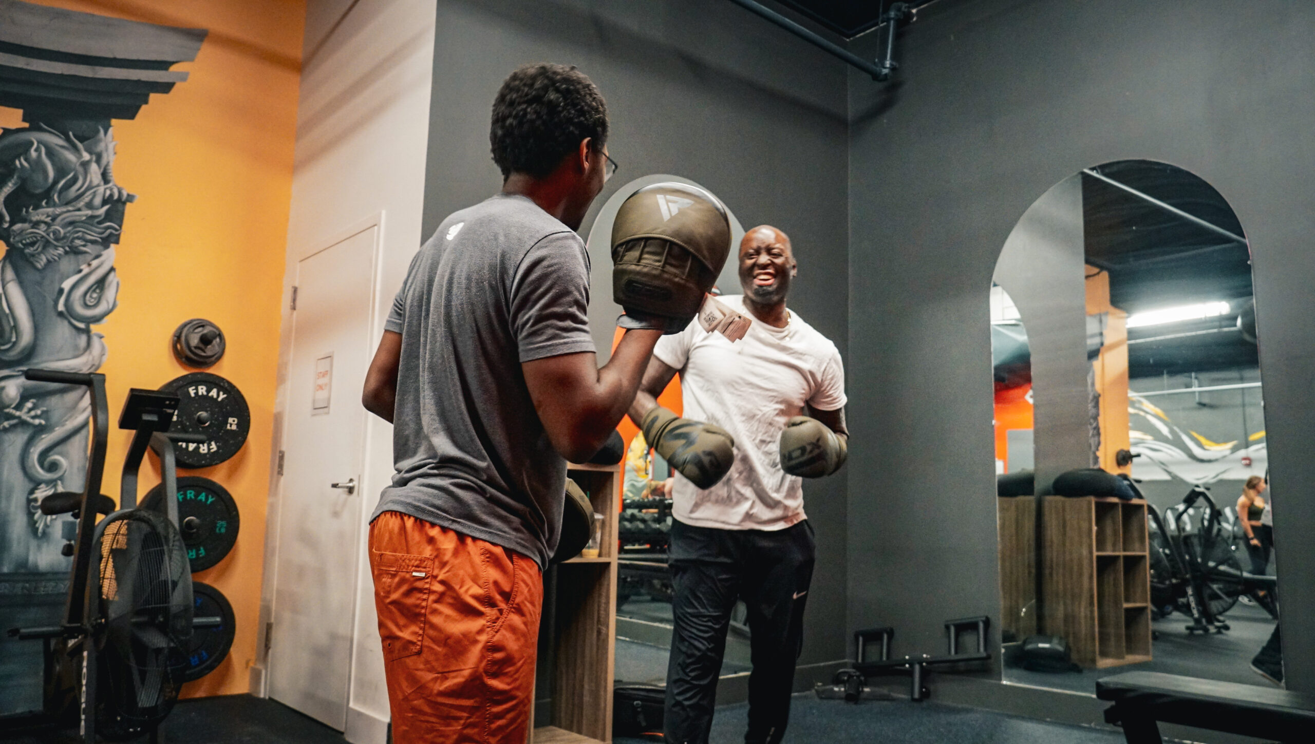 Coach Sergio with his back to the camera, holding boxing mitts while guiding a client wearing boxing gloves at AMPT Fit. The client is smiling and focused during the training session, highlighting the engaging and supportive atmosphere.