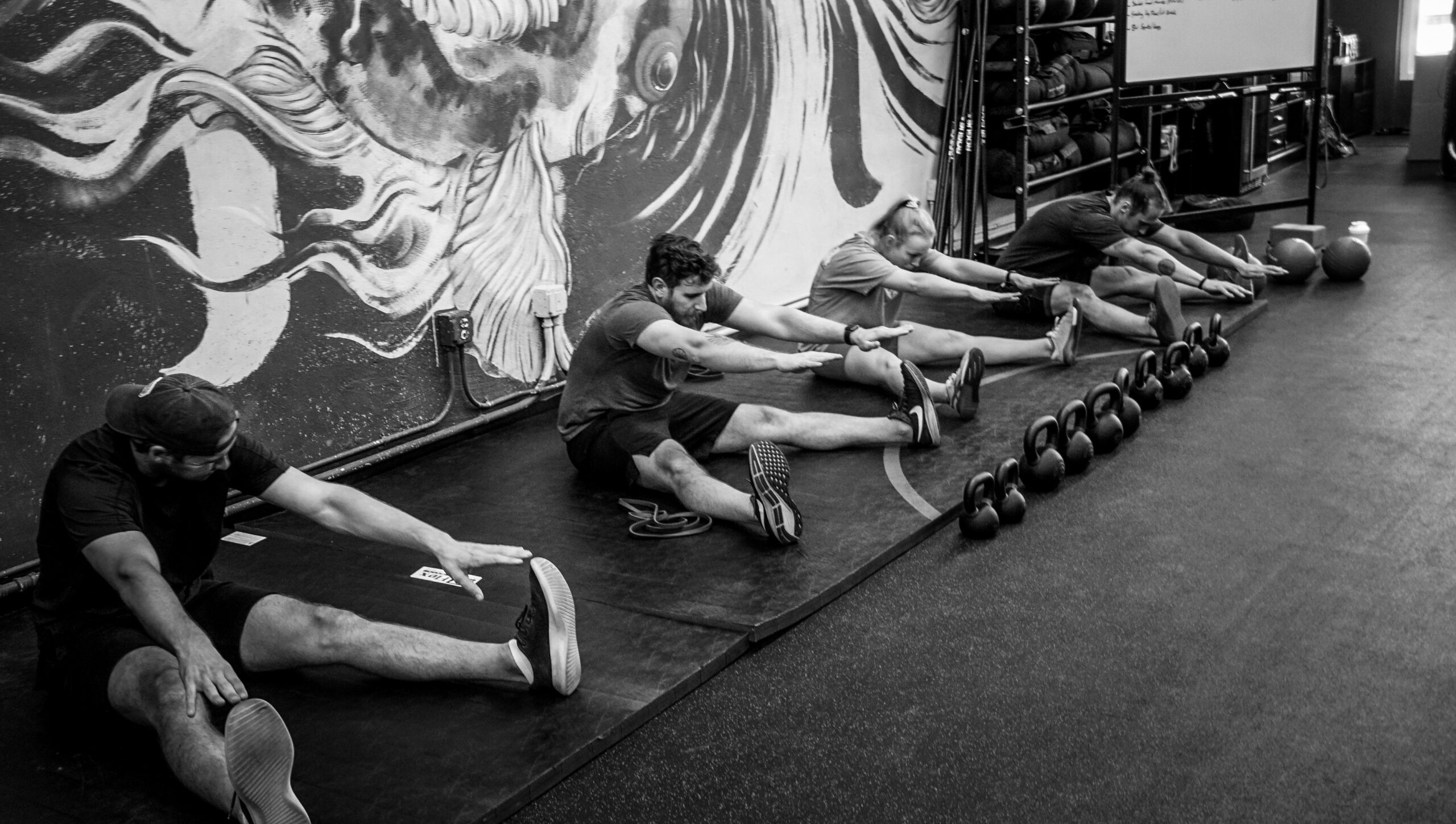 Black and white image of a group of people sitting on the floor and stretching with kettlebells lined up beside them at AMPT Fit gym. Participants extend their arms towards their feet, focusing on flexibility and mobility.