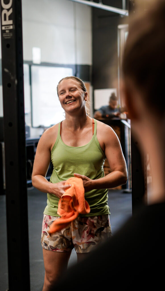 Katherine smiling and holding a towel at AMPT Fit after a workout session, dressed in a green tank top and patterned shorts. She exudes positivity and strength, reflecting her dedication to fitness and well-being.