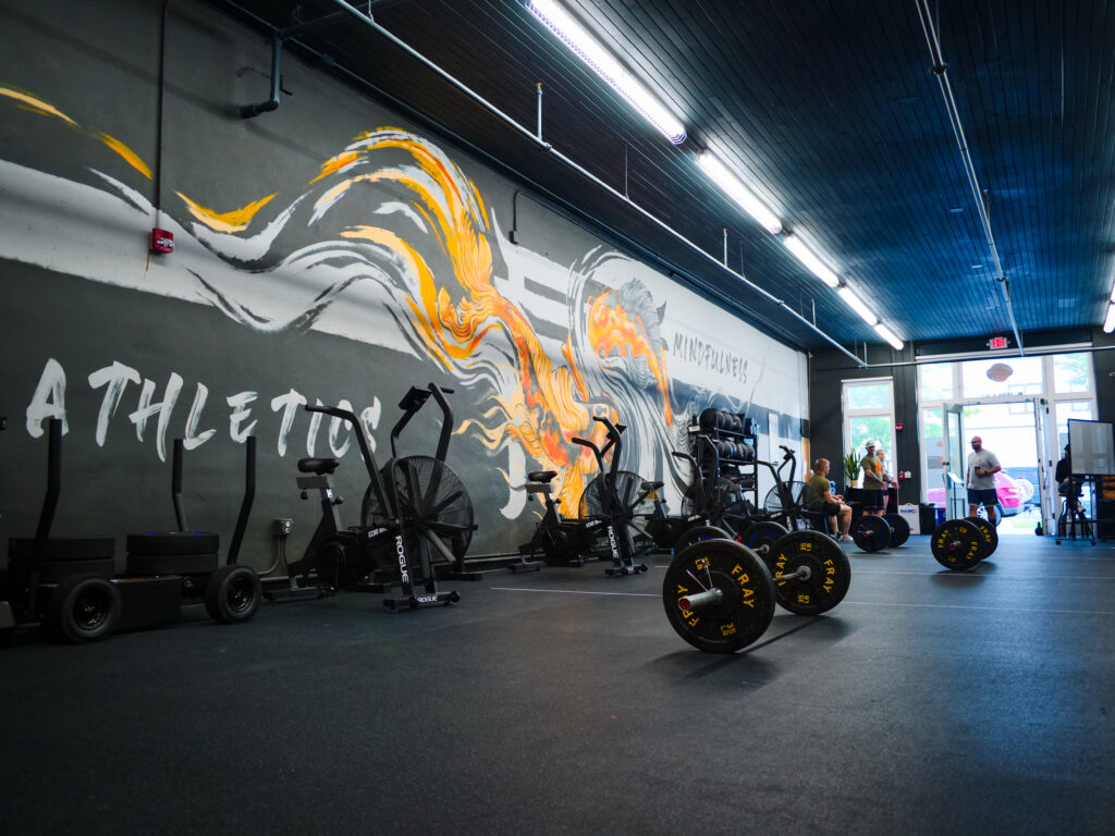 Interior view of AMPT Fit gym, featuring exercise equipment like stationary bikes, weights, and sleds against a backdrop of a vibrant wall mural with the words 'Athletics' and 'Mindfulness'. The spacious, modern facility is well-lit, emphasizing a professional yet welcoming environment for fitness enthusiasts.