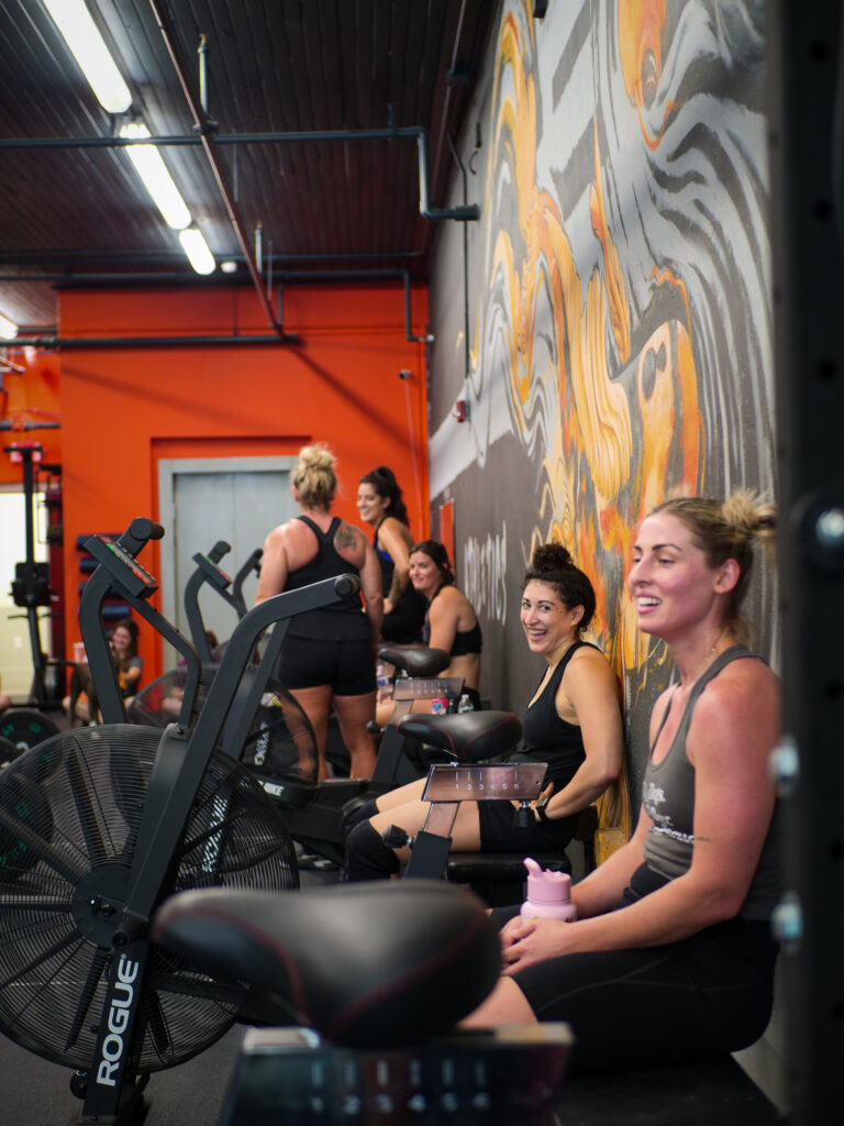 Group of women relaxing and smiling after a workout session at AMPT Fit, seated on exercise bikes and talking in front of a vibrant wall mural. The camaraderie and positive energy reflect the gym’s supportive community and holistic fitness approach.